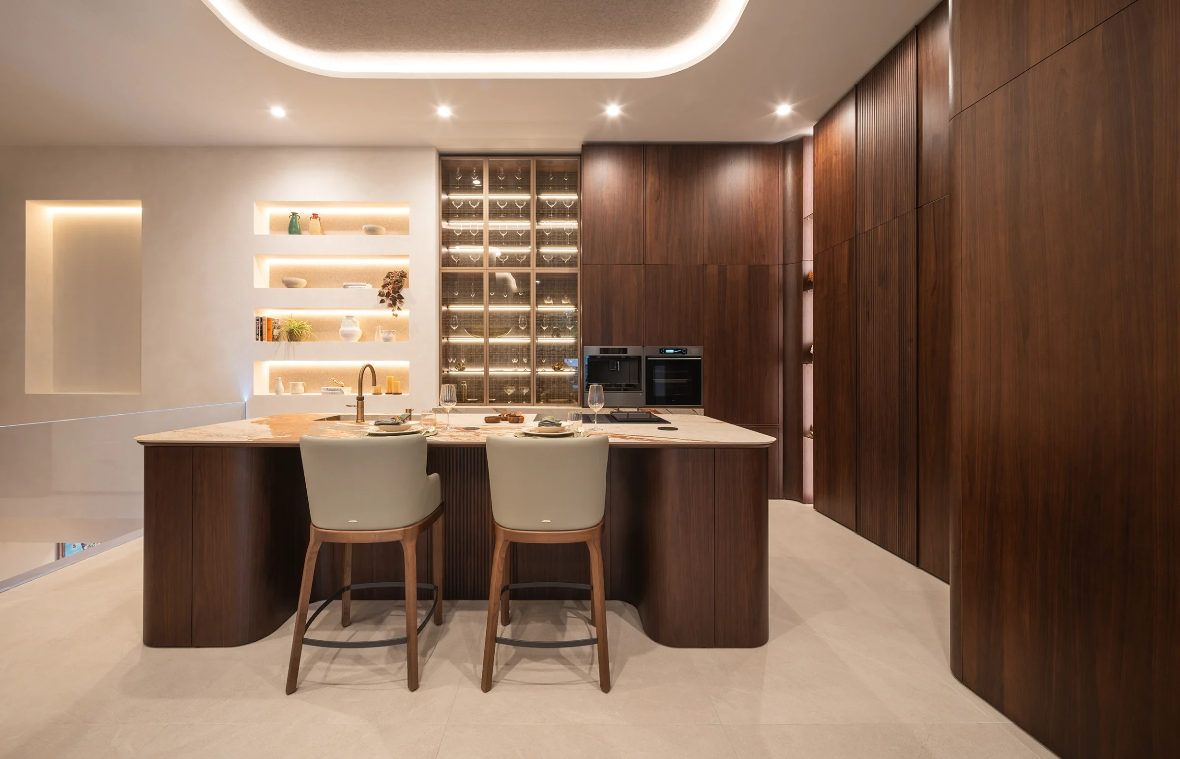 Cocina TERRA de frente: isla curva de madera con encimera de piedra y dos taburetes, vitrinas iluminadas y columnas de madera a la derecha